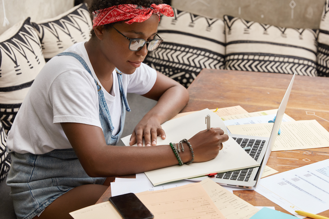 Sideways shot of black busy female journalist studies papers for writing article, uses online issue on laptop computer, sits at wooden desk, writes something down, wears denimm sarafan and spectacles